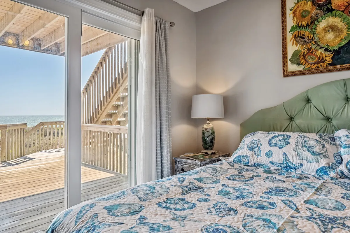A master bedroom with a bed dressed in a seashell-patterned quilt, a large window framing expansive ocean views, a decorative lamp, and framed sunflower artwork. The ambiance is peaceful and inviting, ideal for restful nights and rejuvenating mornings.