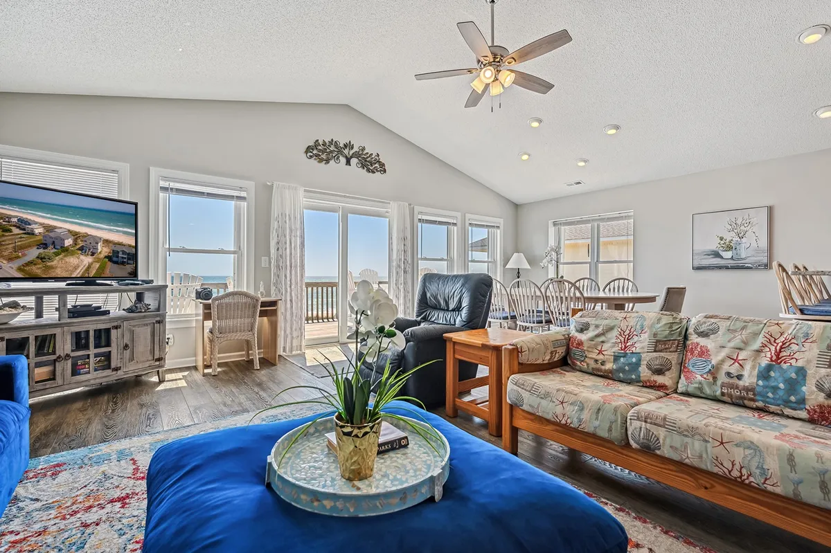 A bright and inviting living area with a colorful couch, coffee table, and large windows offering a clear view of the ocean. The TV also displays a beach scene, reinforcing the indoor-outdoor connection.