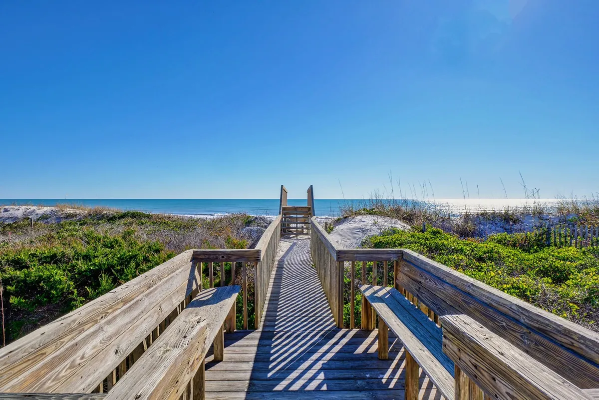 This image features a tranquil exterior scene with a wooden boardwalk leading through dune vegetation toward the beach, with the ocean visible in the background. The soft lighting suggests either early morning or late evening, inviting thoughts of peaceful strolls or starting the day with yoga on the sand.
