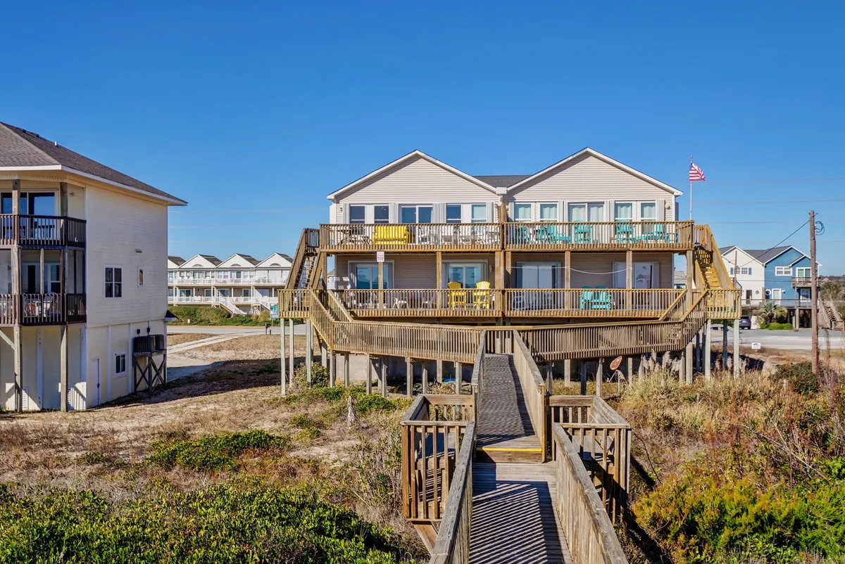 This image shows the exterior of the property with a welcoming wooden deck, yellow chairs for lounging, a walkway leading up to the house, and the American flag flying. The beach is nearby, creating a clear sense of proximity to the ocean and outdoor gathering spaces.