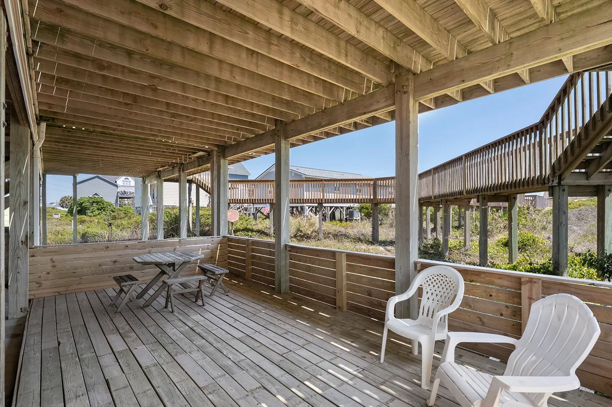 A wooden oceanfront deck furnished with a folding table and white chairs, overlooking the surrounding beach landscape. The view invites relaxation, outdoor dining, and socializing, with the beach just steps away.
