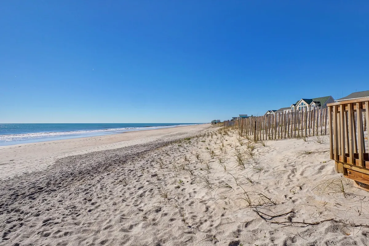 A panoramic view of a sandy beach with ocean waves rolling in, bordered by a wooden deck and fence. The space is open and inviting, perfect for sunbathing, walking, and beachcombing, with clear potential for children’s play.