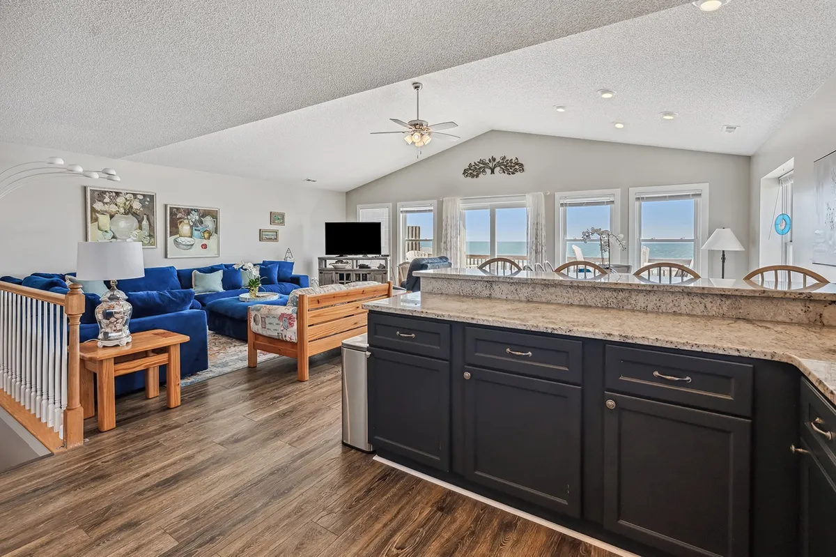 An inviting interior space featuring a blue sofa set, granite kitchen countertops, and expansive windows offering panoramic ocean views. The space is designed for family gatherings, cooking, and enjoying sunsets together.