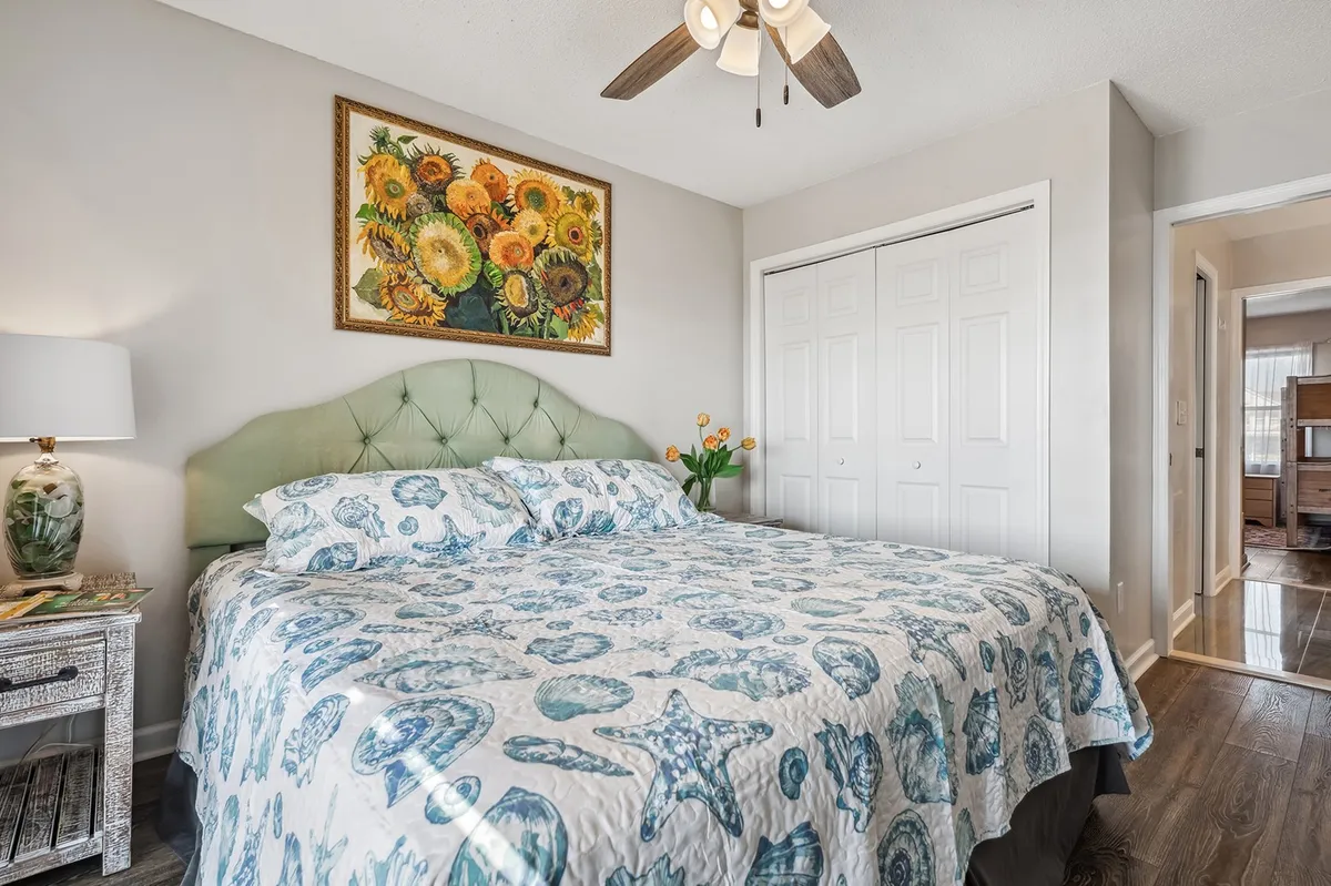 A tranquil bedroom featuring a bed with patterned bedding, a green headboard, a floral painting above the bed, and a nightstand with a lamp. Morning light softly fills the space, creating a peaceful environment ideal for relaxation and restful sleep.