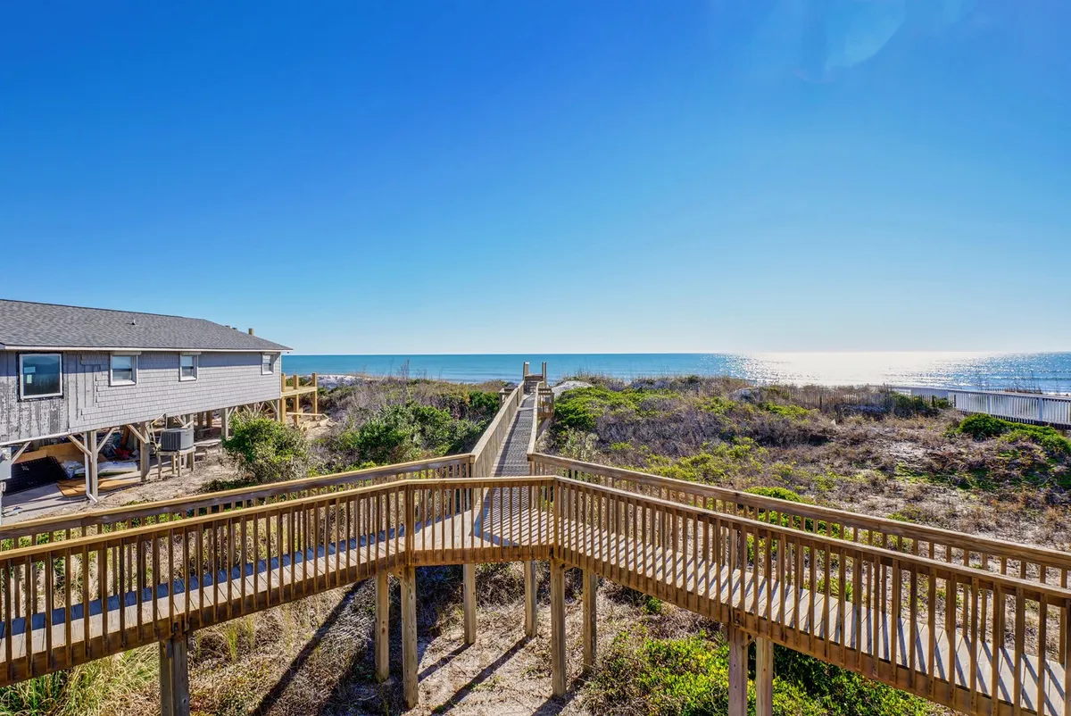 A serene scene of a wooden walkway meandering through coastal vegetation towards a sandy beach and the ocean beyond, with the house set in the background. The setting feels private yet inviting for group adventures.