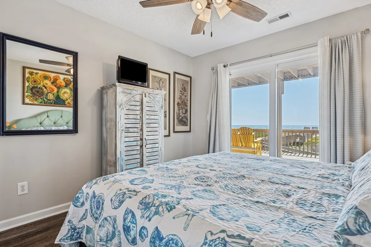 A serene bedroom featuring a bed with a blue and white patterned quilt, sliding glass doors that open directly to a private deck, and colorful floral artwork on the wall. Natural light pours in, creating a tranquil atmosphere that highlights the room’s connection to the outdoors.