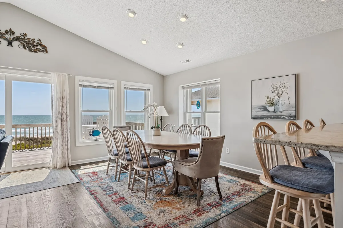 This photo features a dining table surrounded by bar stools, positioned in front of large windows that open onto an ocean view. The area includes a rug and is set for casual dining or entertaining, with sunlight streaming in.