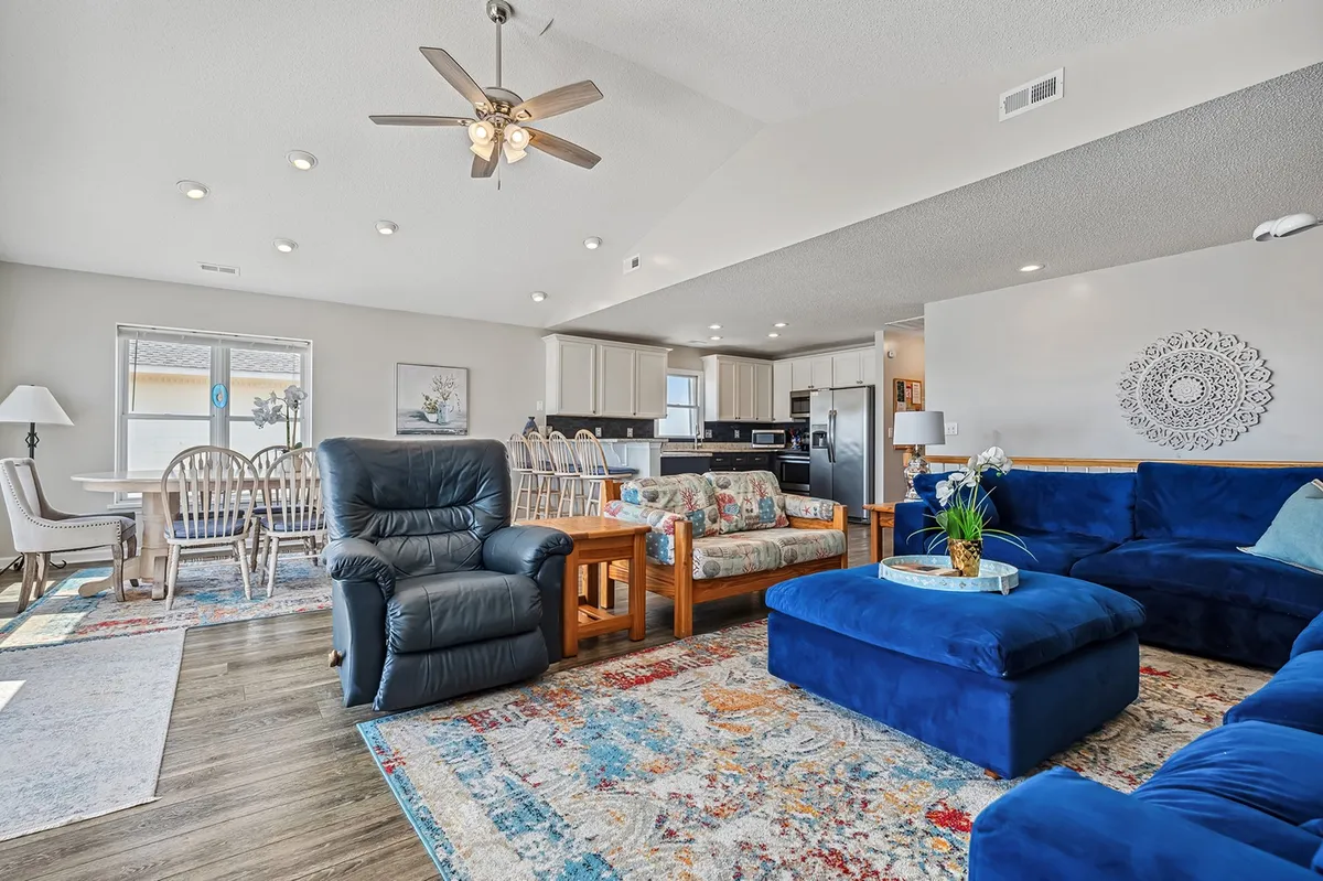 An inviting and spacious living/dining area with a blue sectional sofa, a dining table, colorful area rug, and a ceiling fan. The layout encourages family gatherings, conversations, and relaxation after beach outings.