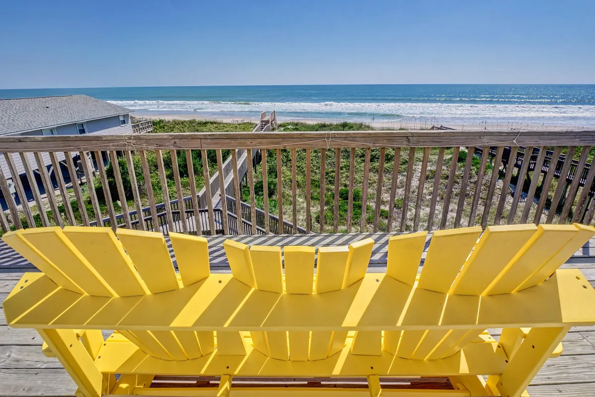 A pair of yellow Adirondack chairs face out over a wooden deck railing toward the sparkling ocean. The scene suggests relaxing moments, dolphin watching, and enjoying panoramic ocean views from an elevated vantage point.