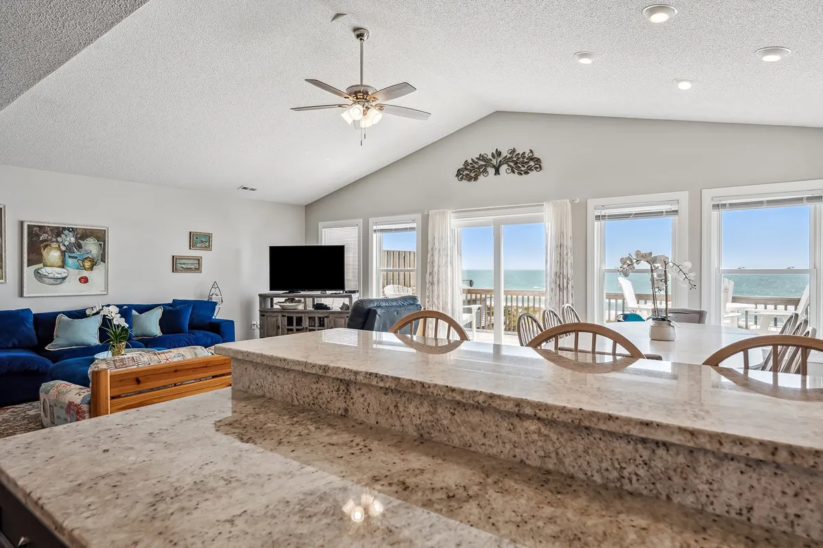 This image captures an expansive open-concept living space featuring a blue sectional sofa, large windows that frame a sweeping ocean view, a granite kitchen island, and a TV unit. The area is bright, airy, and filled with natural light, emphasizing both relaxation and social gathering opportunities.